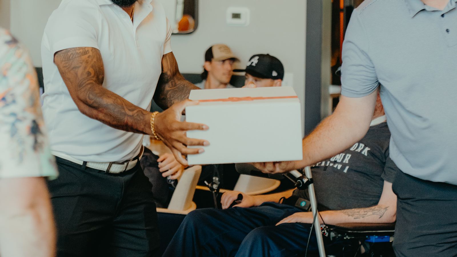 A golfer in a white polo hands a prize box across to another player during the awards at the 2026 Celebrity Golf Classic. Tim Shaw is between them in his wheelchair.